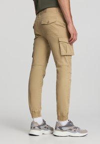 Beżowe spodnie cargo z elastycznymi mankietami, z bocznymi kieszeniami i kieszeniami z tyłu. Noszone z szarymi butami z siateczki i beżowymi sneakerami.