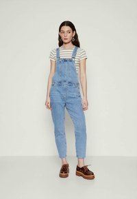Jonge vrouw in gestreepte t-shirt, lichtblauwe denim overalls, bruine platform schoenen en hartvormige oorbellen, staand tegen een effen achtergrond.