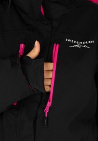 Black softshell jacket with pink zippers and accents. Features a logo on the chest, and a thumb hole in the sleeve for added comfort.