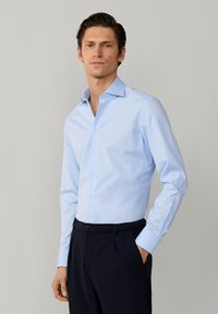 Mann trägt hellblaues Hemd und dunkle Hose, steht mit einer Hand in der Tasche vor schlichtem grauem Hintergrund.