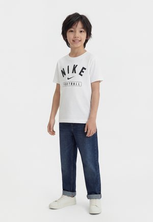 Fant stoji, oblečen v belo Nike nogometno majico, modre kavbojke z manšetami in bele superge, se smehlja v kamero.