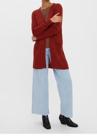 Cardigan lavorato a maglia in rosso mattone con apertura frontale, motivo testurizzato e tasche a toppa, abbinato a jeans a gamba larga azzurri e stivaletti neri.