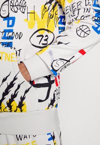 Sweat-shirt blanc avec des imprimés colorés style graffiti comprenant des ballons de basket, le numéro 73, des flammes et des formes abstraites, avec une main dans la poche avant.