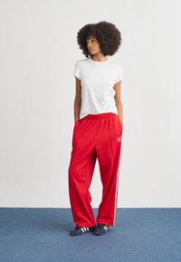 Calças desportivas vermelhas com riscas laterais brancas, combinadas com uma t-shirt branca simples e sapatilhas azuis-marinho, apresentando cintura elástica.