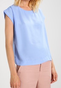 Bluzka z krótkim rękawem w jasnoniebieskim kolorze, wykonana z gładkiego materiału. Posiada okrągły dekolt i prosty dół, zestawiona z różowymi spodniami.