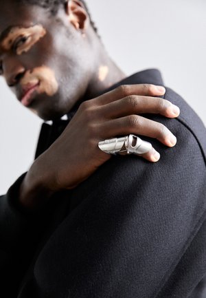 Bague en métal au design sculptural, dotée d'une finition argentée polie, de découpes et d'une forme angulaire, portée sur une main sur un tissu sombre.