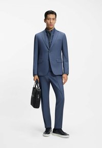 Man draagt een blauw pak met een bijpassend zip-up shirt, houdt een zwarte aktetas vast en draagt zwarte sneakers met witte zolen.