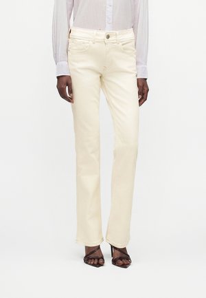 Femme portant un jean crème à jambes droites, une chemise légère rayée boutonnée et des sandales noires à talons hauts et ouverts aux orteils, debout devant un fond blanc.