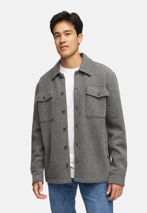 Giacca grigia a coste con colletto, bottoni frontali, due tasche sul petto e maniche lunghe, indossata sopra una camicia bianca.