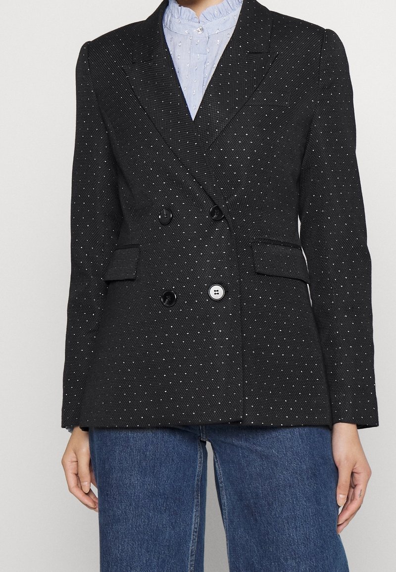 Blazer noir à double boutonnage avec de petits pois blancs, porté sur un chemisier bleu clair texturé avec un col à volants et un jean en denim bleu.