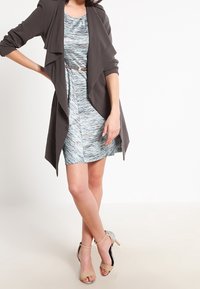 Cardigan gris drapé sur une robe bleue texturée avec un motif subtil, cintrée à la taille avec une ceinture. Sandales à talons beiges.