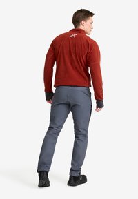 Man som står, iklädd en röd fleecejacka, grå fritidsbyxor med sidfickor och svarta vandringsskor, vänd bort från kameran på en vit bakgrund.