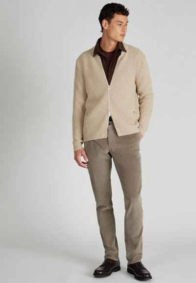 Beige geribbelde cardigan met een rits, gedragen boven een donkerbruine overhemden met kraag, gecombineerd met lichtbruine gestructureerde broek en donkere schoenen.