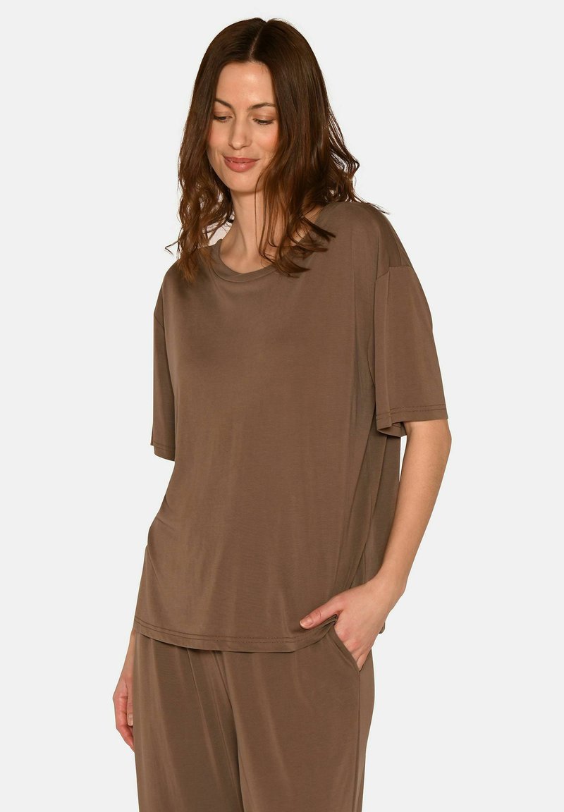 Mujer con cabello castaño hasta los hombros, vistiendo un top marrón de manga corta y pantalones a juego, posando con una mano en el bolsillo.