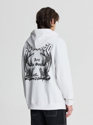 Persona che indossa una felpa bianca con cappuccio con illustrazioni nere di angeli e testo in stile gotico sul retro, in piedi davanti a uno sfondo chiaro e uniforme.