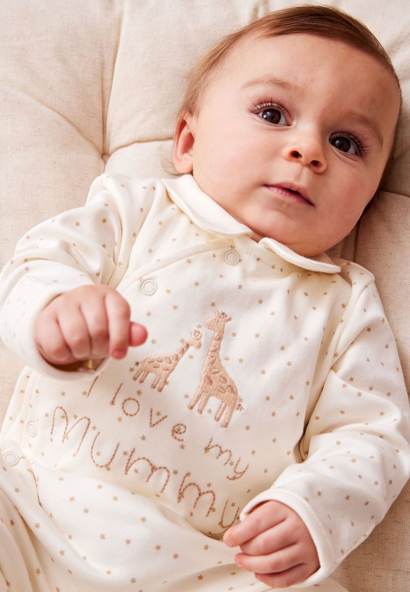 Bambino sdraiato su un cuscino beige che indossa una tutina bianca con piccole stelle, la scritta "I love my mummy" e un ricamo di giraffa.