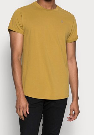 Mand iført en karrygul t-shirt med korte ærmer og sorte bukser, der står mod en lys baggrund.