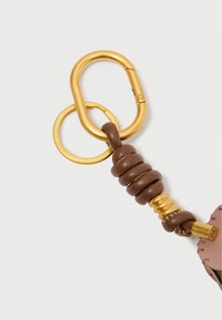 Brahns ādas atslēgu piekariņš ar saritinātu dizainu, zelta krāsas aparatūra ar ovālu un apaļu gredzenu, kā arī dekoratīvu šuvju akcentu.