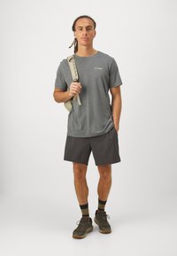 Tricou gri cu mânecă scurtă și un mic logo, asociat cu pantaloni scurți închiși la culoare, șosete cu dungi negre și maro, și pantofi de drumeție gri. Ține un rucsac bej.