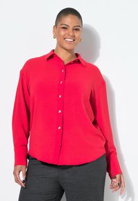 Smiling person with short hair wears a red button-up shirt and gray pants, posing against a white background.