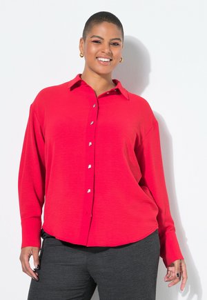 Una persona sorridente con capelli corti indossa una camicia rossa a bottoni e pantaloni grigi, posando su uno sfondo bianco.