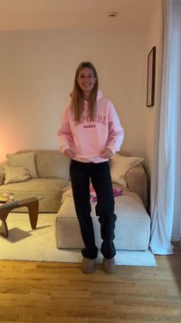 Ροζ φούτερ με το κείμενο "Hawaii", μαύροι παντελόνια και μπεζ παντόφλες. Μακριά μαλλιά ακατάστατα, στέκεται σε δωμάτιο με καναπέ και τραπεζάκι του καφέ.