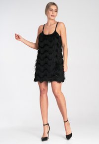 Schwarzes Fransenkleid mit wellenförmiger Textur und dünnen Trägern, kombiniert mit schwarzen Stiletto-Absätzen. Minimalistisches Design, knielang.