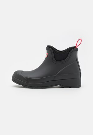 Black waterproof ankle boot made of rubber, featuring a textured surface, flat sole, and a red pull tab at the back for easy wearing.