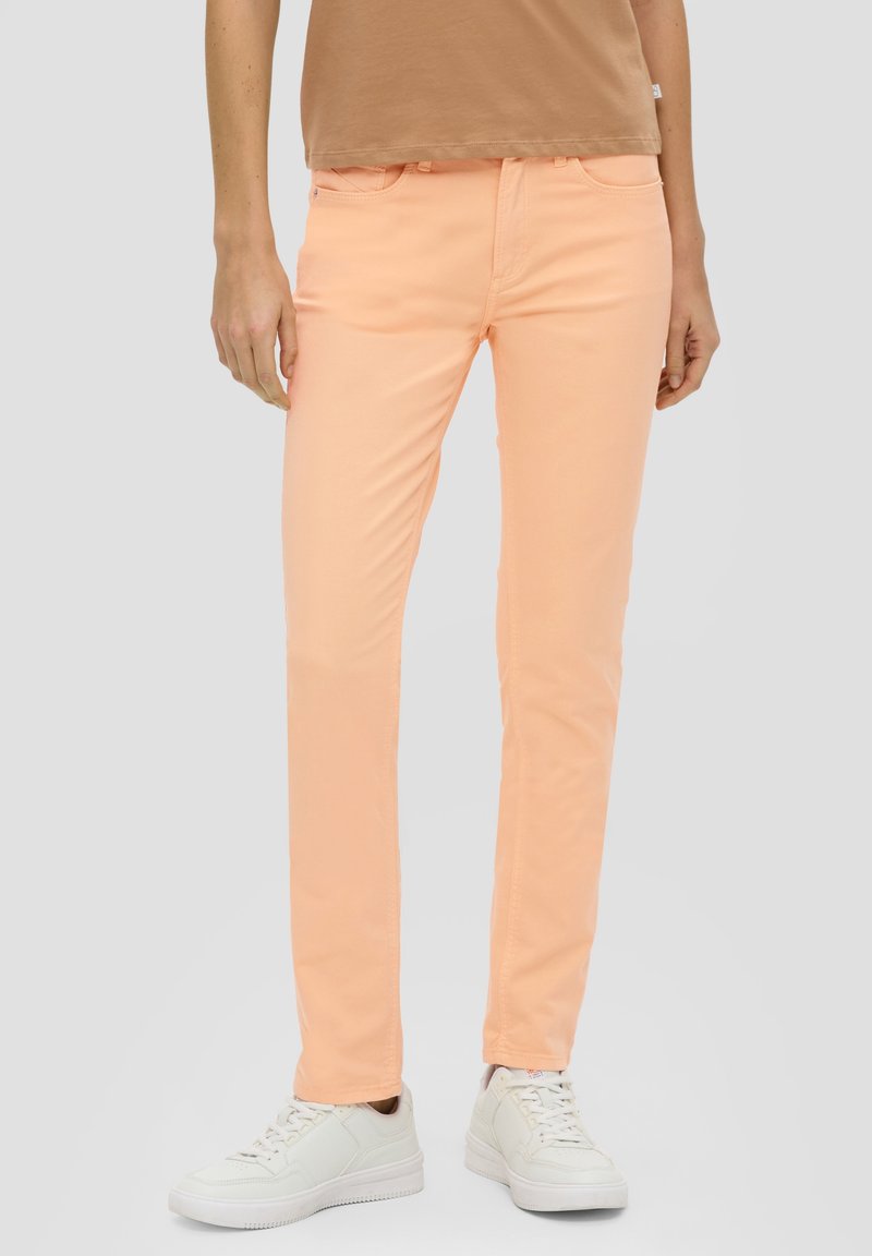 Persona con pantaloni aderenti color pesca, sneakers bianchi e un top corto beige su uno sfondo chiaro uniforme.