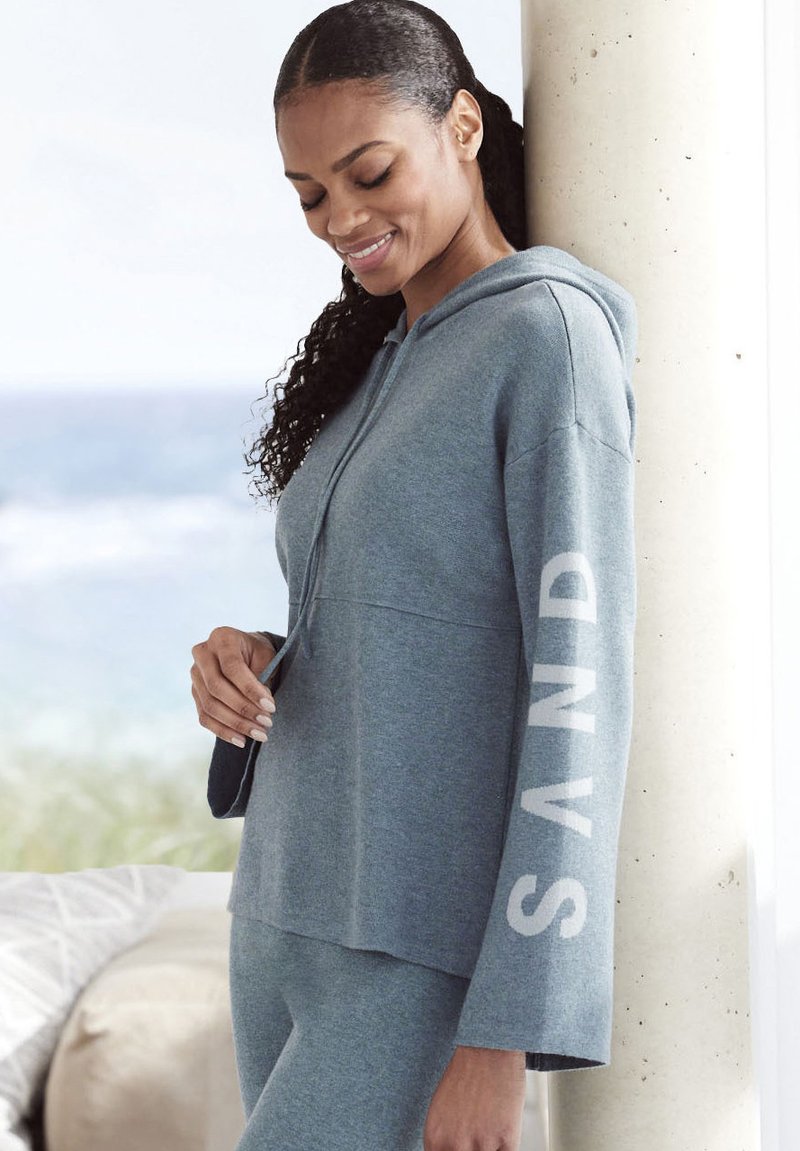 Felpa grigia con cappuccio e scritta "SAND" stampata in bianco sulla manica. Tessuto morbido, vestibilità comoda e maniche lunghe. Abbinata a pantaloni coordinati.