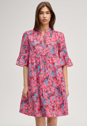 Kobieta ubrana w różową sukienkę w kwiaty sięgającą kolan, z niebieskimi liśćmi, falbaniastymi krótkimi rękawami i wiązaniem z przodu przy dekolcie, stojąca na jednolitym tle.