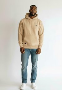 Homme debout portant un sweat à capuche beige, un jean bleu clair et des baskets blanches sur un fond clair uniforme.
