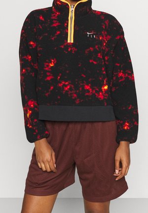Persoon draagt een zwart-rood patroon fleece trui met gele rits en bordeauxrode mesh short, staand tegen een grijze achtergrond.