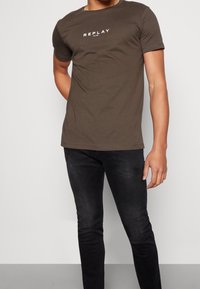 Olivově zelené tričko s krátkým rukávem, na němž je bílý nápis "REPLAY". V kombinaci s těsnými černými džíny. Bavlněný materiál, hladká textura.