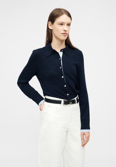 Γυναίκα που φοράει καφέ μπλε ζακέτα με κουμπιά πάνω από ανοιχτό γαλάζιο πουκάμισο, λευκό παντελόνι ψηλής μέσης και μαύρη ζώνη, στέκεται με το ένα χέρι στην τσέπη.