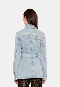 Giubbotto di jeans azzurro chiaro con vita a cintura e ricamo "LIUJO" sulla schiena. Presenta maniche lunghe e un colletto classico.