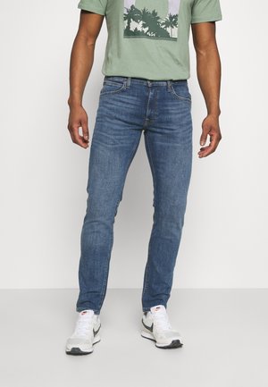 Mies, joka pukeutunut slim-fit-mallisiin sinisiin farkkuihin, valkoisiin Nike-tennareihin ja vihreään t-paitaan, jossa on musta palmukuosi valkoisella taustalla.