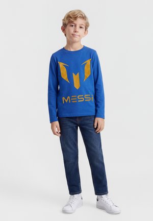 Lyshåret dreng stående iført en blå langærmet skjorte med guldtekst og logo "MESSI", mørke jeans og hvide sneakers på hvid baggrund.