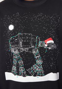 Felpa nera con un design a tema nevoso che presenta un AT-AT illuminato decorato con luci e un cappello di Babbo Natale contro uno sfondo stellato.