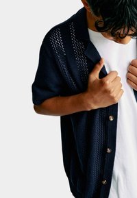 Cardigan corto de manga corta en color navy con textura de punto, que presenta acentos de tejido abierto y un cuello redondeado. Cierre con botones plateados en la parte frontal.