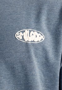 Blå meleret stof t-shirt med et buet broderet hvidt "smilodox"-logo på venstre brystområde. Blødt, tekstureret materiale.
