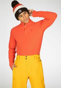 Heldere oranje lange mouwtop met een halve rits, gecombineerd met hoogzichtbare gele broek met twee zwarte knopen op de taille. Gebreide muts met een pompon.