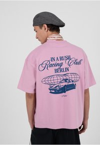 Roosa lühikeste varrukatega t-särk, millel sinine tekst "IN A RUSH Racing Club BERLÍN" ja graafika autost koos maakeraga, lahtise lõikega.