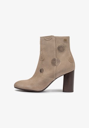 Botín beige de ante con tacón cuadrado, puntera redondeada, parches circulares decorativos brillantes y pequeño logo dorado cerca del borde superior.