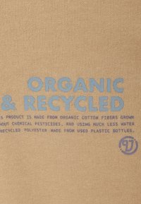 Tissu beige avec une texture tricotée visible comportant du texte bleu et violet. Les détails mettent en avant des matériaux en coton biologique et en polyester recyclé.