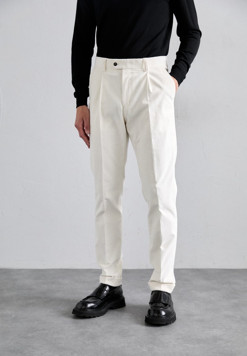 Pessoa vestindo calças tailandesas em creme com pregas e sapatos loafers pretos, de pé contra uma parede e chão cinza claro.