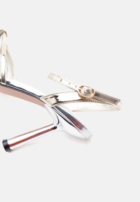 Ασημί πέδιλο με ψηλό τακούνι, λεπτά λουράκια και χρυσή αγκράφα, με λαμπερό μεταλλικό φινίρισμα και λεπτό τακούνι με μαύρη άκρη.