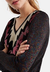 Pull à sequins avec un col en V, présentant des motifs en zigzag multicolores rose, or et noir sur un fond sombre et texturé.