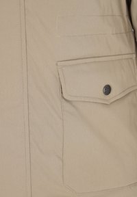 Acercamiento de tela beige que muestra un bolsillo con solapa y un botón metálico en una prenda de vestir.