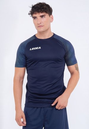 Uomo che indossa una maglietta da ginnastica blu navy con maniche decorate e logo "LEGEA", in piedi con una mano vicino alla vita.